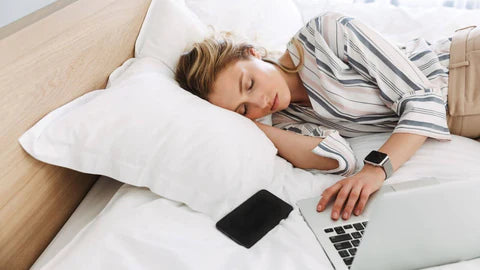 woman laying down in bed with her head on a pillow. It appears she has fallen asleep while working. Her laptop and cell phone are in the bed with her, her hand on the laptop and the cell phone right next to the laptop.