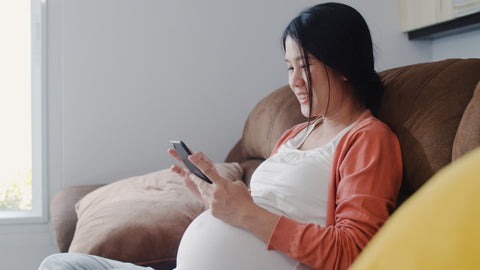 young Asian woman sitting on couch, pregnant, with cell phone in her two hands right up against her pregnant bump