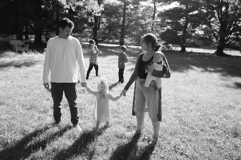 Matthew and Chanel are holding Lyra's hands and walking towards the camera. Chanel is looking at Matthew and holding newborn daughter, Lily. Sons, DJ and Emmett are in the background, sword fighting with sticks. They are outside in their backyard and the behind them are mature pine trees.