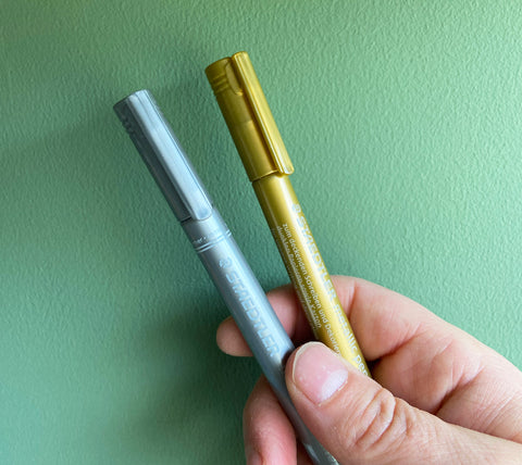 Gold and silver marker pens in front of a green background.