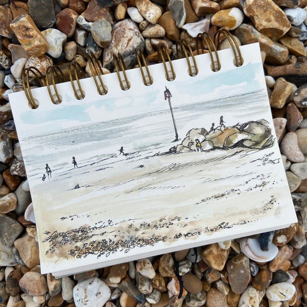 Ringbound sketchbook on a pebble beach with a watercolour painting on the open page