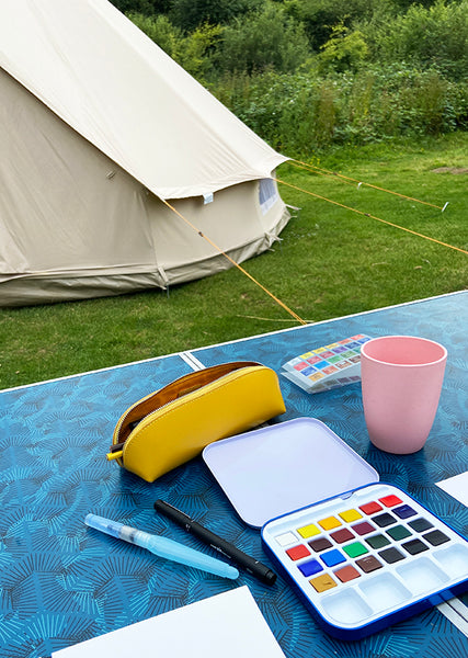 Travel watercolour paints and mustard coloured pencil case on a camping table in front of a bell tent