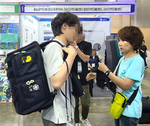 A customer guy purchasing a large backpack with braille patches on it. A Feeldom staff is wearing a bright green mini sling bag and helping the man put on the backpack