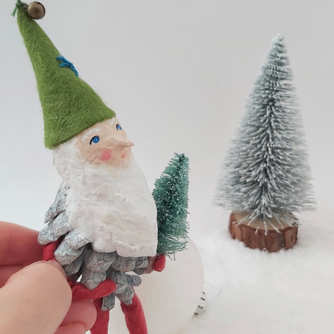 spun cotton pine cone elf ornament, held by hand against a white background with bottle brush tree in the distance