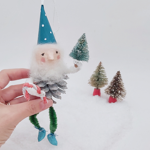 Hand holding a spun cotton pine cone elf against a white background with two bottle brush trees in the distance. 