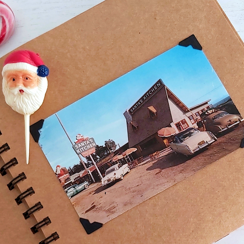 A scrapbook opened to a vintage Christmas postcard from Santa's Kitchen, California. A vintage Santa ornament sits above.