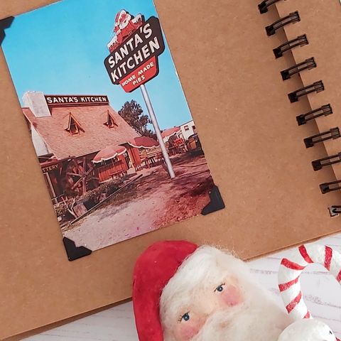 A scrapbook opened to a vintage Christmas postcard from Santa's Kitchen, California. A spun cotton Santa peeks from below.