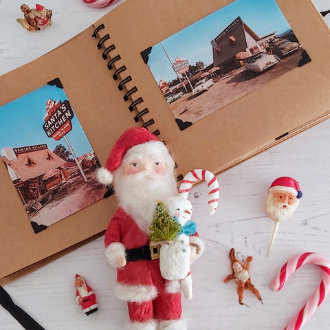 A scrapbook opened to two vintage Christmas postcards from Santa's Kitchen, California. A spun cotton Santa sits in front along with candy canes and vintage Santa decorations.