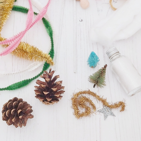 2 pine cones, bump chenille pipe cleaners, bottle brush trees, bottled glitter, cotton, and tinsel laying on a white wood table background