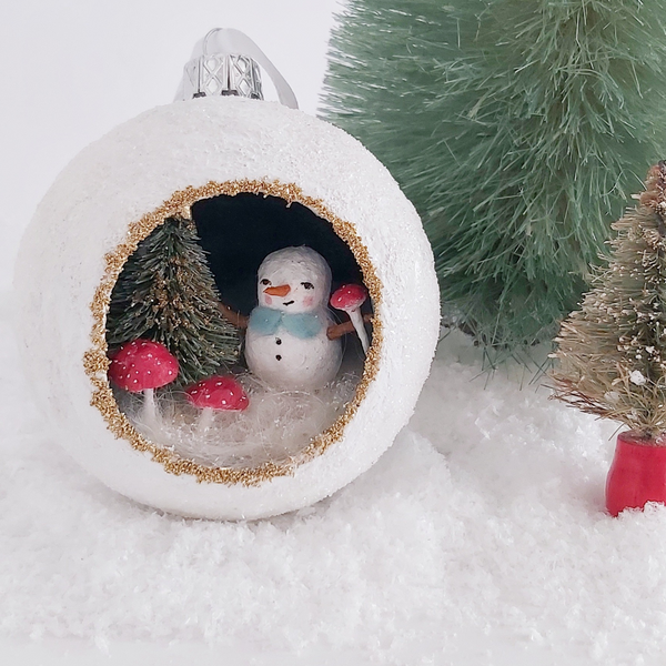 a spun cotton snowman inside of a paper mache diorama ornament, next to a green bottlebrush tree