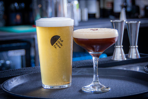 A full pint of fresh Full Circle Brew Co Lager, sat on a Taproom Bar Top on a serving tray alongside an Espresso Martini served in a small cocktail glass.