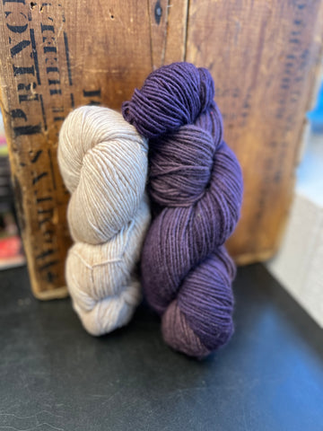 two skeins of yarn, one white and one purple, standing on end against a wooden box on a dark surface