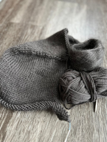 A partially knitted hood in a cool brown shade laying on a dark wood floor