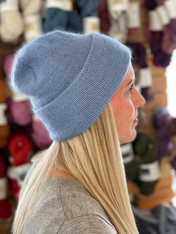 A person with long blonde hair wearing a blue fuzzy hat standing in fron of skeins of yarn