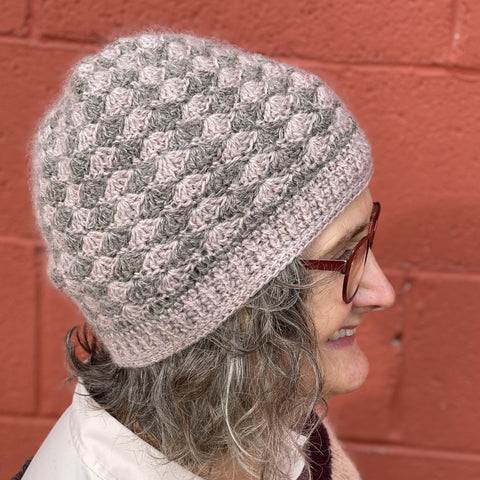 Photo of a pink and grey crochet hat, shown in profile on a person wearing glasses
