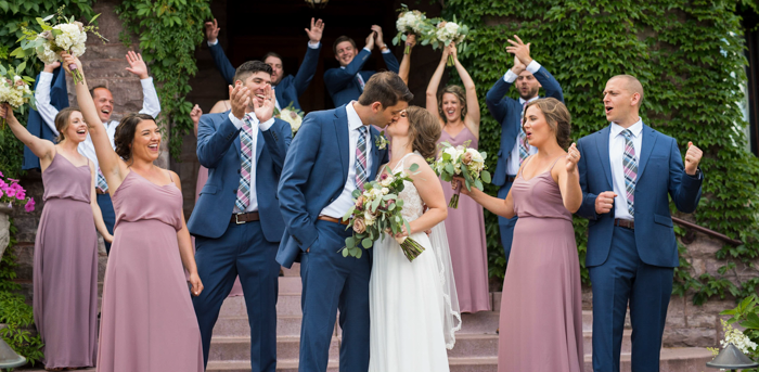 A bride and groom kiss while surrounded by their bridal party in front of an ivy covered building