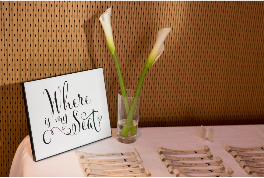 A sign saying "where is my seat" is next two a vase with two calla lilies and place cards