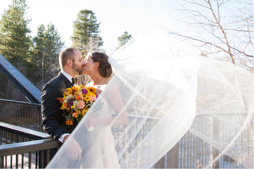A bride's veil blows in the wind while she kisses her groom on a deck
