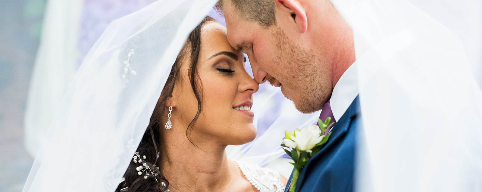 A bride and groom are underneath the veil with their noses touching and their eye closed.