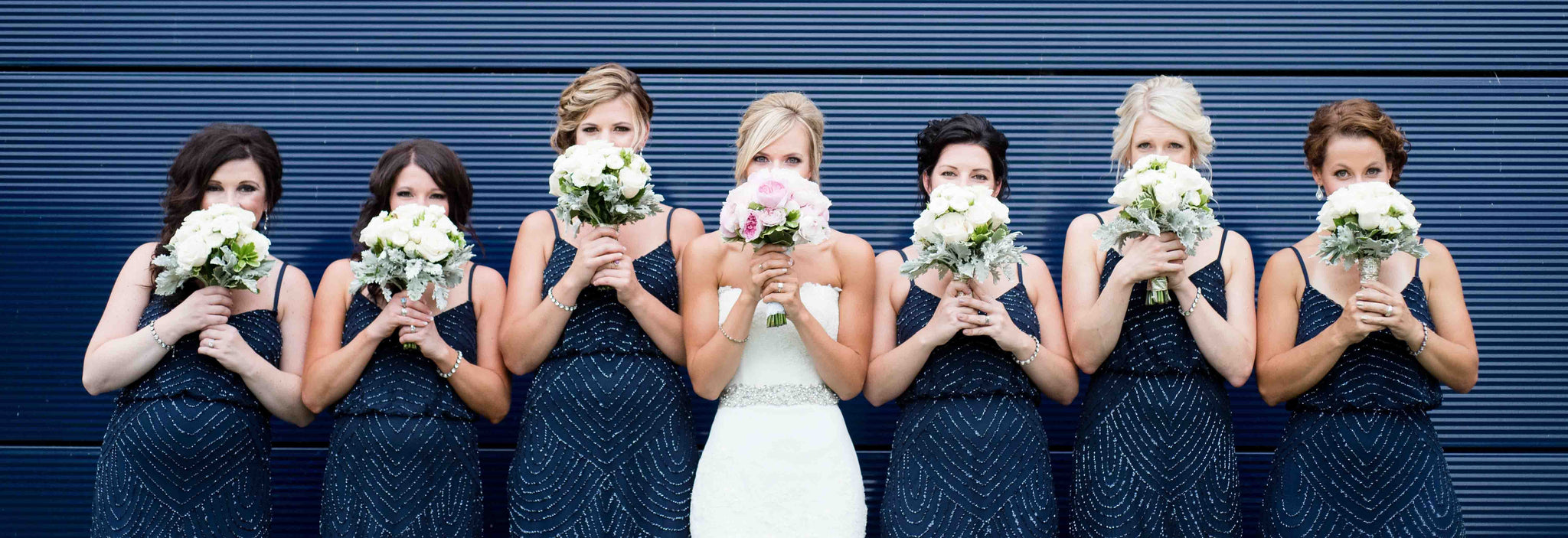 A bride and her bridesmaids, wearing blue dresses, hold their bouquets in front of their faces