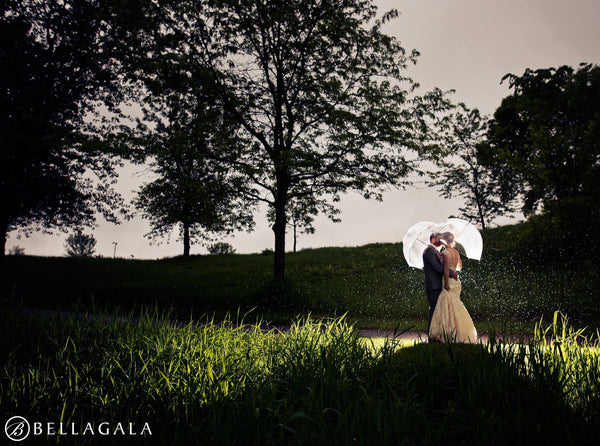 A couple kissing underneath umbrellas while backlit in a garden. A wide shot showing the dramatic gray of the sky to create contrast among the dark trees.