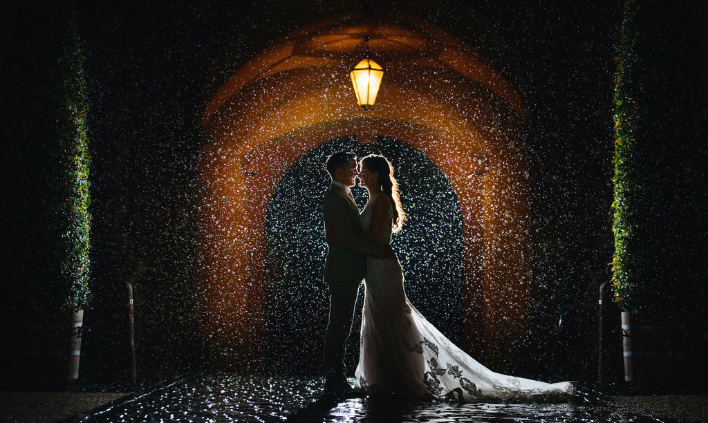 A silhouette of the couple is backlit from a light underneath an archway. Rain falls, creating a twinkling effect from the light source.