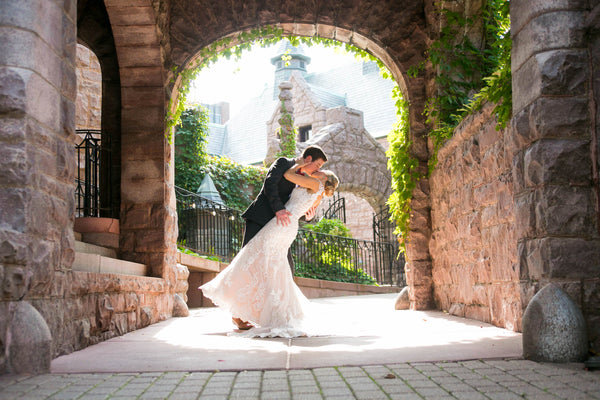 A groom is dip kissing his bride under the brick archway of the Van Dusen Mansion.