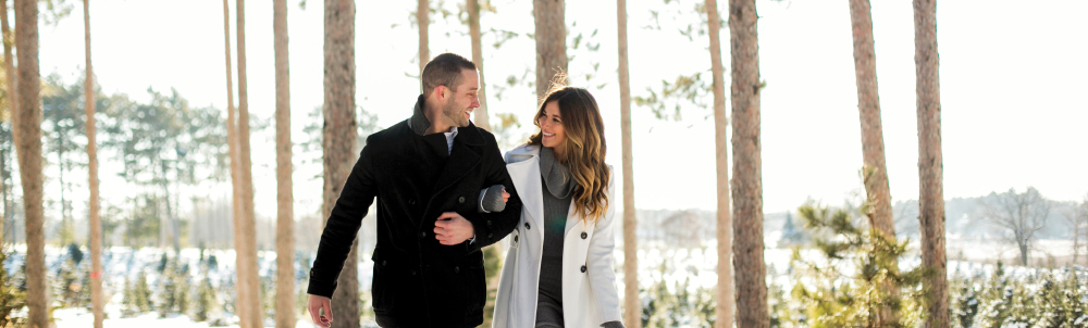 A man in a black coat and a woman in a white coat walk arm in arm through trees.