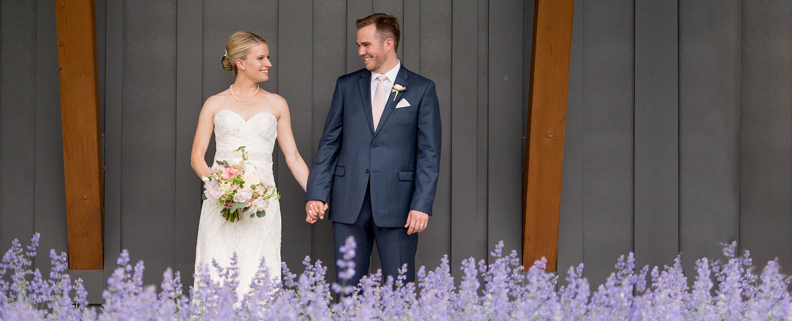 A bride and groom stand behind a row of lilacs, holding hands and smiling at each other