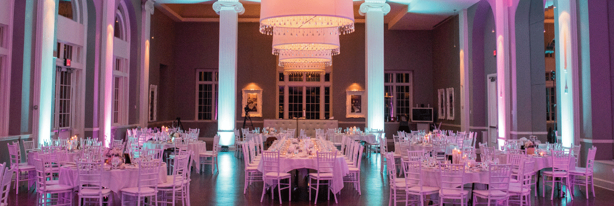 A reception space with white columns, white tables and white chairs with alternating purple and blue uplighting on the columns