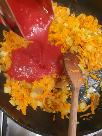 tomato passata being added to a pan with chopped onions and grated carrots