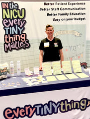 NICU nurse Trish Ringley RN standing in Synova NICU Leadership Forum Exhibit hall behind a table displaying NICU journals and NICU milestone cards for hospital purchase. Standing in front of banner with the words "better patient experience, better staff communication, better family education, easy on your budget"