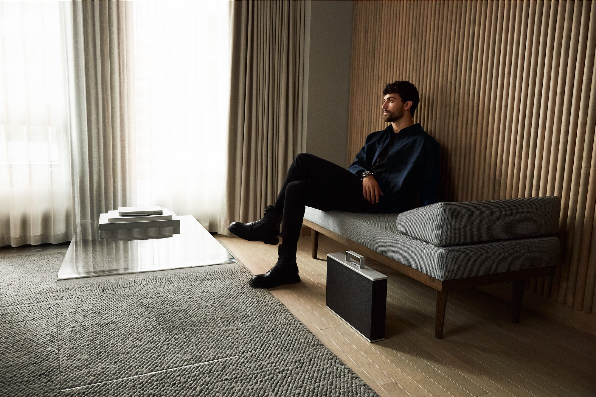 Wide shot of young man with Mackenzie watch case wearing luxury suit sitting on a grey couch in a room with modern interior design. Art books are propped on a sleek glass table. The watch case made from aluminum and carbon fiber , premium young bull leather and soft Alcantara is standing on the floor.