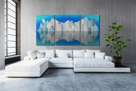 A spacious modern living room with large windows, featuring a three-piece blue-toned wall art depicting mountain reflections in water, a large sectional white sofa, and a potted green plant to the right. The painting is Kenai Fjords National Park by Topher Straus