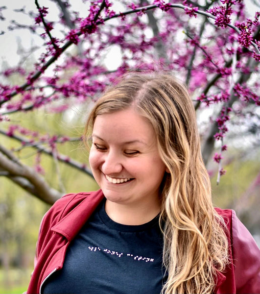 A close up of Frankie Ann with a huge smile and looking away from the camera. She is wearing a black t-shirt from Aille Design with custom braille beadwork that reads "elle jones casting" in silver Swarovski Crystal Pearls. Frankie Ann paired her t-shirt with a red leather jacket. She is standing outside in front of a Cherry Blossom tree in full bloom.