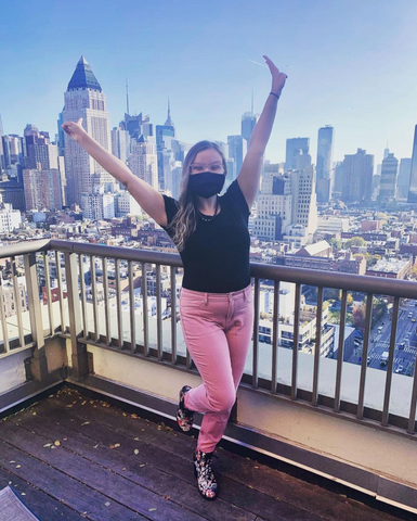 Frankie Ann is filled with joy, standing in front of the NYC skyline with her arms up in the air. She is wearing clear glasses, a black mask, black t-shirt, pink pants, and floral lace up boots.  