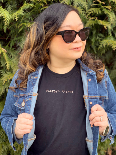 Anne wearing Aille Design Custom Braille T-Shirt “purpose in view”. The t-shirt is black and the braille is created with silver Swarovski Crystal Pealrs. Anne styled her custom braille t-shirt with a denim jacket and black sunglasses. She is standing in front of beautiful greenery and looking away from the camera.