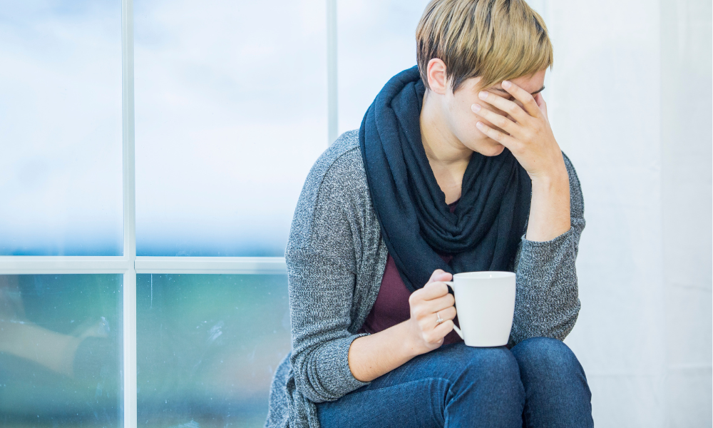 Person, feeling low, holding their head by the window, wearing a jumper and scarf