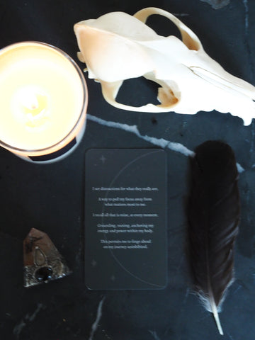 moon oracle card with feather, crystal wand, lit soy candle, and animal skull on black background