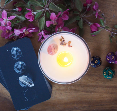 top view of crystal infused gemini zodiac candle with moon mantra oracle cards on wooden background with pink flowers and astrology dice