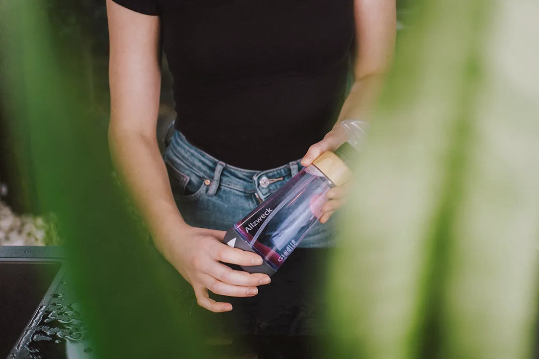 A young woman is standing at the kitchen worktop and is holding a WingGuard cleaning bottle filled with water and a cleaning agent tab.