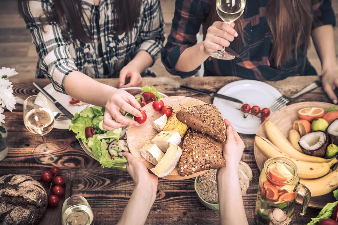 Young people sit together at a large wooden table with various dishes and hand each other a cheese platter.