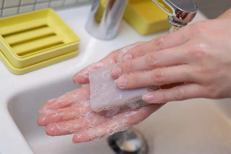Eingeschäumte Frauenhände halten Lavendelseife. Im Hintergrund ein Waschbecken mit fließendem Wasser und eine gelbe Seifenschale.