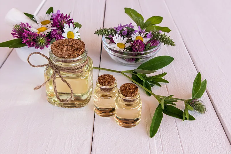 Natürliche Kosmetik in Glasflaschen aus einem weißen Holztisch umgeben von Kamille, Gänseblümchen, Klee und Minze.