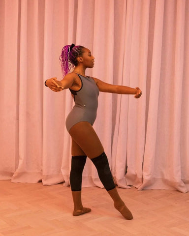 Ruth Essel founder of Pointe Black, wearing a grey leotard stretching on the floor of a ballet studio