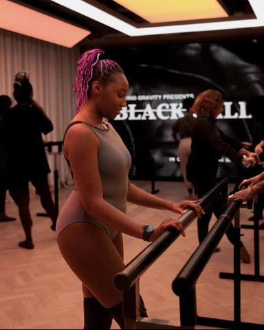 Ruth Essel, Founder of Pointe Black. A Black Ballerina wearing a grey leotard dancing at the barre in a black ballet class