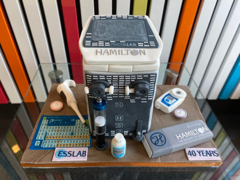 A cake shaped like a Hamilton ML600 Dilutor/Dispenser surrounded by other items in ESSLAB's portfolio, such as syringes and syringe filters.