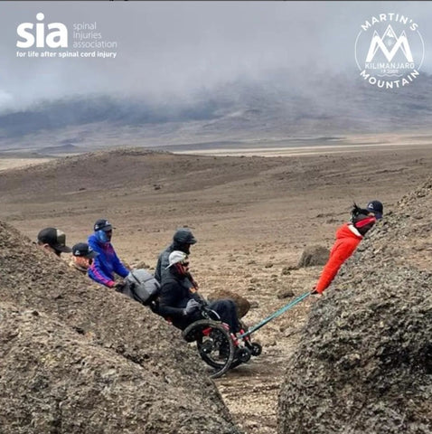 photo shows a man in a wheelchair being helped by porters, in a barren landscape