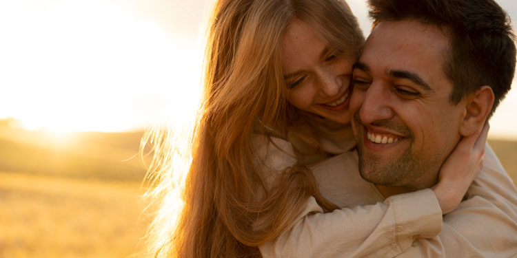 Woman riding her man piggyback and hugging him; both smiling; meadow in background