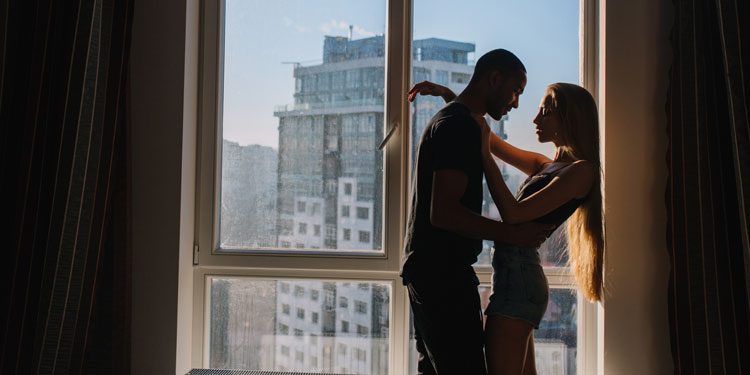 a photo of a young couple playfully standing in front of a window and flirting with each other